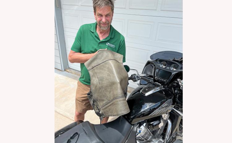 Tim Schanno stands next to his motorcycle parked in the driveway of his Hot Springs home while he holds up the vest he was wearing when he was shot in the back. With his left hand, Shanno presses through the vest to show the bullet hole in the leather. Photo by Brett Nachtigall/ FRC Herald-Star
