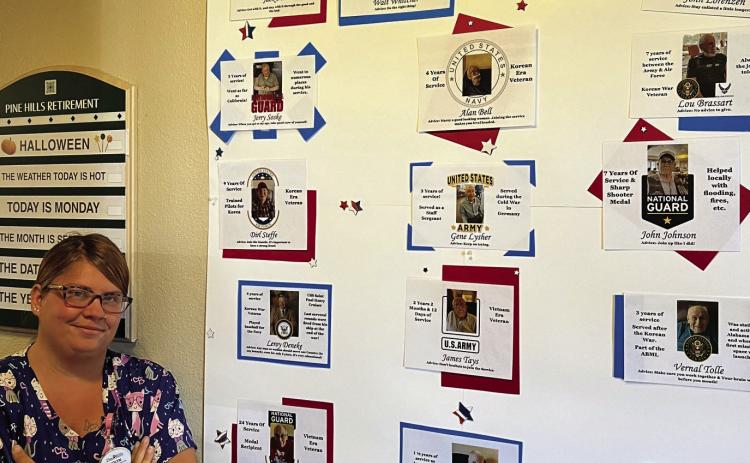 Jolyn Harder, Pine Hills Retirement Community Lead Med Aide, stands by the wall of heroes she created at Pine Hills honoring residents who served our great country. Photo by Charity Maness/Fall River County Herald Star